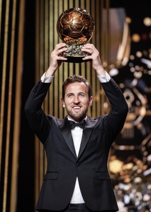 Harry Kane in a black tuxedo holding up the Ballon d'Or trophy on a golden stage.
