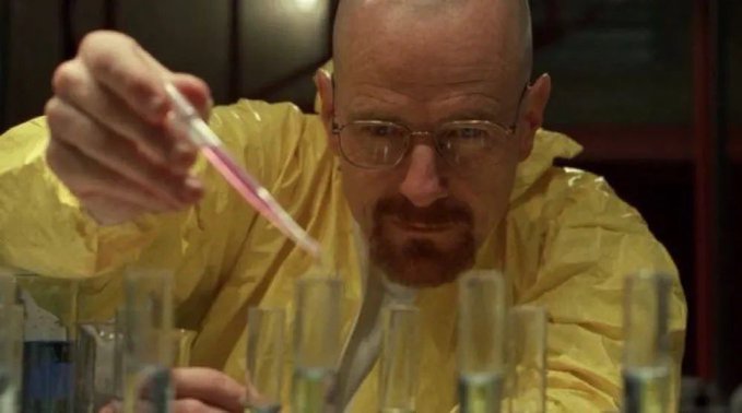 A still from Breaking Bad showing Walter White in a yellow hazmat suit and glasses, holding a pipette over several test tubes in a makeshift laboratory.