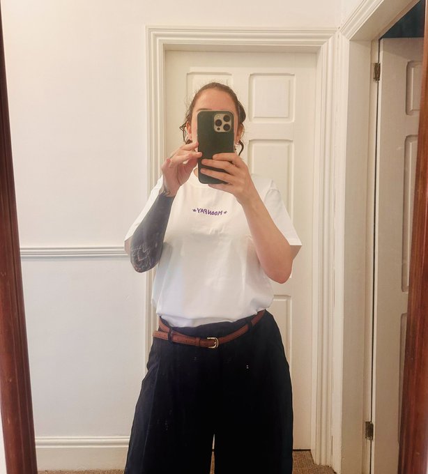 A person stands in front of a mirror taking a mirror selfie, wearing a white t-shirt with blue text reading "*YAH00M*", dark trousers, and a brown belt.