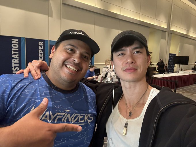 Two smiling young men pose together at what appears to be an indoor convention or tournament event, one wearing a blue 'WAYFINDERS' jersey and cap giving a shaka hand sign, the other in a black cap and hoodie with white earbuds.