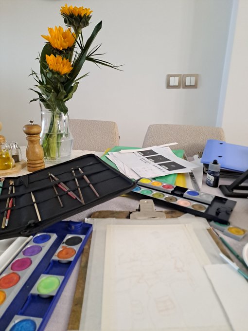 An artist's workspace featuring sunflowers in a glass vase, a wooden pepper grinder, watercolor paint palettes, brushes in a black case, and a painting in progress on an easel.