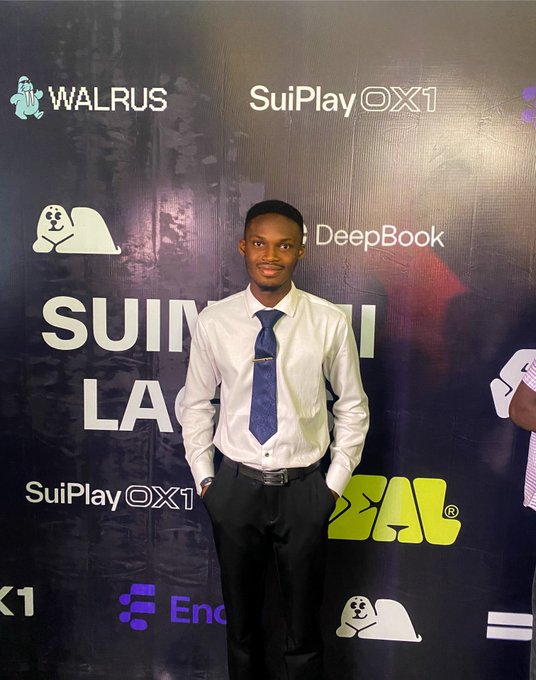 A man wearing a white shirt, dark tie, and black pants posing in front of a backdrop with logos including 'WALRUS', 'SuiPlay 0X1', 'DeepBook', 'REAL', and 'SUIM... LA...'.