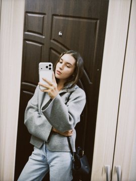 A young woman in a gray hoodie takes a mirror selfie with a white smartphone in front of dark brown double doors.
