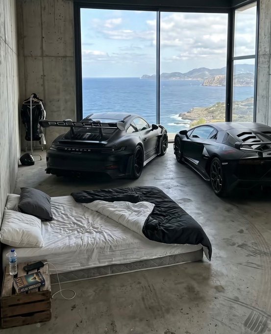 A luxurious industrial-style bedroom features an unmade bed with rumpled white and black bedding in the foreground, with two matte black Porsche sports cars parked inside next to floor-to-ceiling windows overlooking a stunning ocean and coastal cliff view. A small wooden crate serves as a nightstand holding a water bottle and a book, while a clothing rack stands near the concrete wall.