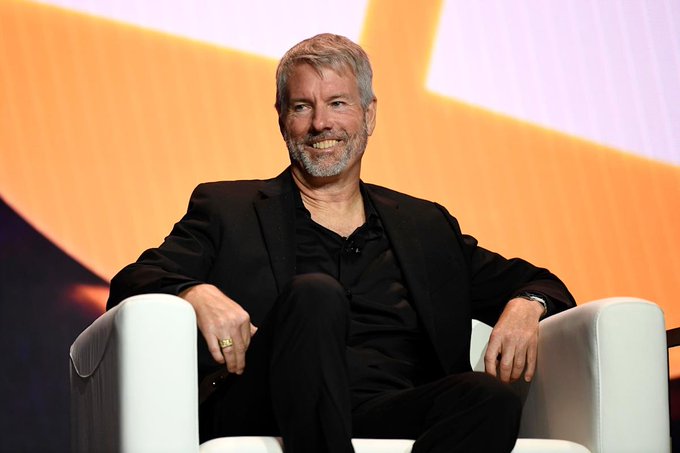 The image features a man sitting in a white chair, smiling at the camera with his hands resting on his knees. The background of the photo shows an orange and purple screen, which appears to be part of a stage or presentation setting. There is also text that reads "Michael Saylor: Confidence in Ethereum has collapsed. Sui has collapsed after being hyped as the 'next Solana.' The rest of the crypto market is fighting over utility." This suggests that Michael Saylor might have been discussing the collapse of Ethereum and the subsequent rise of Bitcoin, which could be related to his own experiences or insights on the matter.