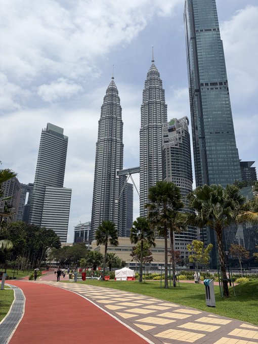 Vertical photo of the Petronas Twin Towers in Kuala Lumpur, Malaysia, viewed from a park featuring a red running track, palm trees, and surrounding skyscrapers under a partly cloudy sky.