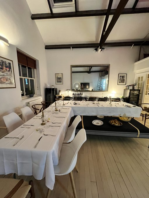A dining room with exposed wooden ceiling beams features tables set with white tablecloths, place settings, and chairs around them, with a large mirror on the wall reflecting the room.