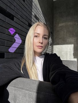 A selfie of a blonde woman wearing a black hoodie, sitting on a grey couch next to a brick wall with a purple digital arrow sticker pointing toward her.