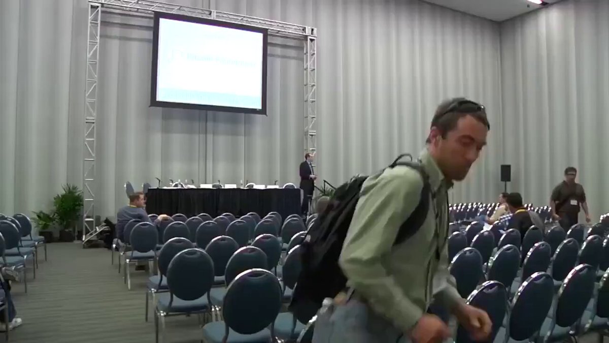 The image depicts a large auditorium filled with rows of chairs, with a presentation area featuring a big screen that displays information related to Bitcoin at $100. A man wearing glasses and a backpack can be seen walking past the seating arrangement towards this demonstration. The focus should be on understanding what is happening in front of him rather than paying attention to his actions or belongings he carries as he moves through the room.