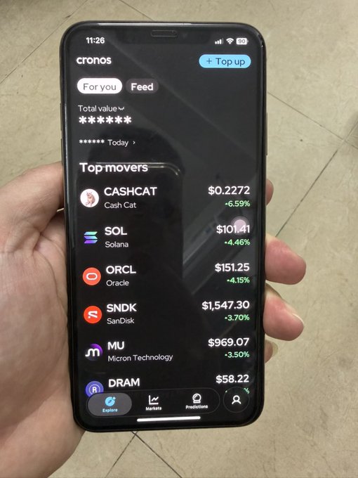 A hand holds a smartphone displaying the 'cronos' crypto app, showing top movers including CASHCAT ($0.2272), SOL ($101.41), ORCL ($151.25), SNDK ($1,547.30), MU ($969.07), and DRAM ($58.22).
