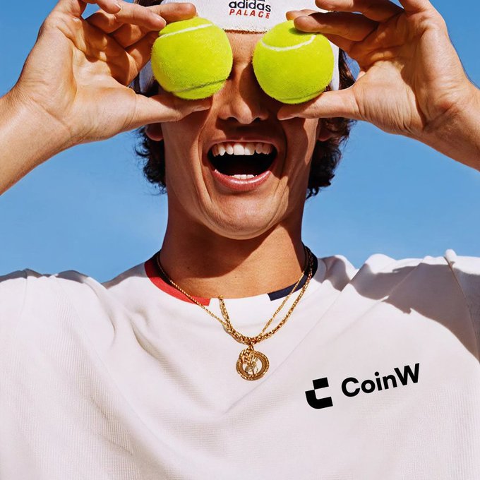 A smiling tennis player holds two tennis balls over his eyes against a blue sky, wearing an Adidas shirt and a CoinW-branded jersey. The “adidas” logo and “CoinW” text are visible, reinforcing the post’s playful comparison between the crypto market and a tennis match.