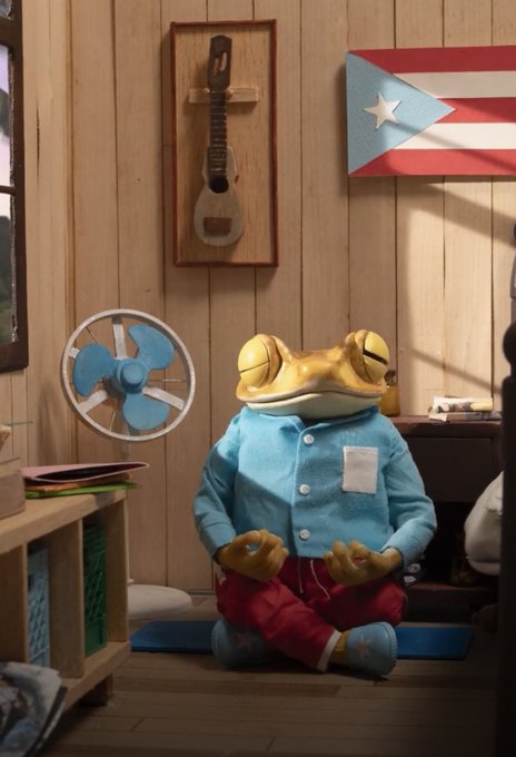 A 3D render of an anthropomorphic frog wearing a blue shirt and red pants, meditating in a yoga pose on a blue mat inside a wooden room decorated with a Puerto Rican flag and a stringed instrument on the wall.