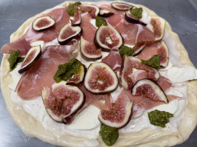 An unbaked pizza topped with fresh fig slices, prosciutto, dollops of creamy white cheese, and green pesto, capturing an experimental fig-season creation in the making.