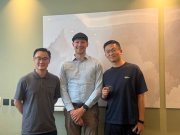 Three men pose for a photo in front of a wall with abstract artwork; the middle man is tall with a beard and blue shirt, flanked by two men in dark polo shirts.