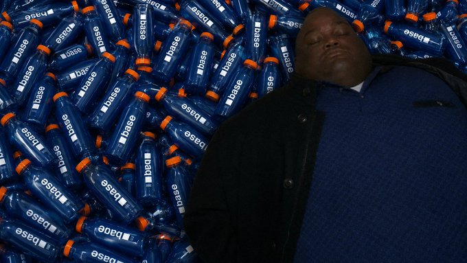 A man sleeps peacefully amid a massive pile of blue "base" branded juice bottles, visually embodying the post's joke about going broke investing in $BASEJUICE, with the contract address "0xb200000000000000000000046390aed221043f01" displayed for potential buyers.