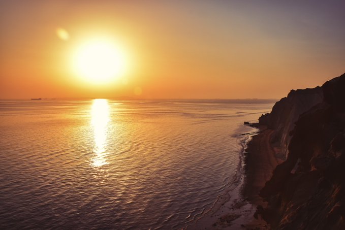 A serene ocean sunset scene with the sun low on the horizon reflecting golden light across calm waters beside dark coastal cliffs.