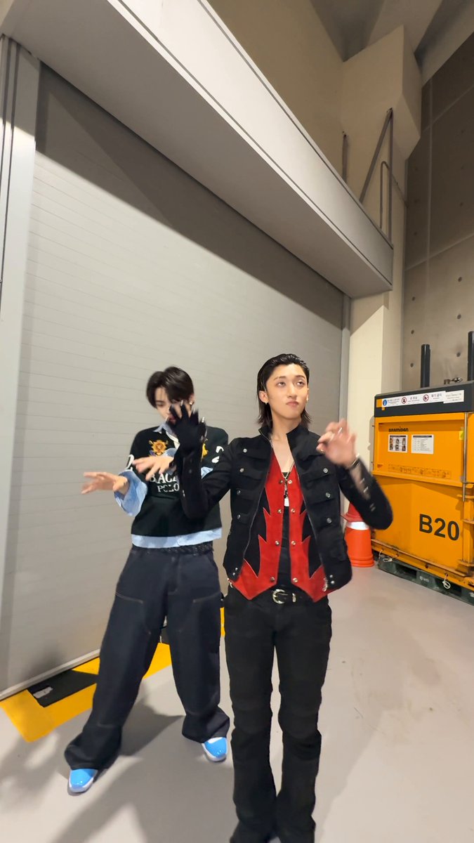 The image features two young men standing next to each other, both wearing black jackets with red accents. One of them has a pair of blue shoes on his feet. The background includes a white wall and an orange dumpster. There are also some text elements in the image that read "#MOYA with #ALPHADRIVEONE #LEO @ALD1_official #LNGSHOT #WOOJIN #4SHOVILLE".