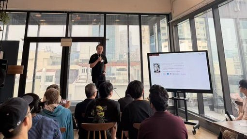 A man stands speaking to a seated audience in a sunlit room with large windows, while a screen displays what appears to be a social media profile feed.