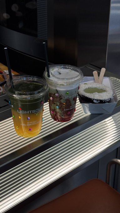 Two iced drinks in clear plastic cups with straws (labeled "LIGHTOUT") and a small white food container sit on a metallic ribbed surface, possibly a drink holder.