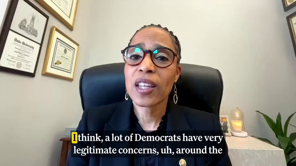 The image shows a woman wearing glasses sitting behind a desk with her hands on her knees while talking into a microphone. The background features several framed pictures, suggesting that she might be in an office or a meeting room. The text "I think, a lot of Democrats have very legitimate concerns around ethics and illicit finance as Congress debates the CLARITY Act." is displayed above the woman's head, indicating her role in the conversation.