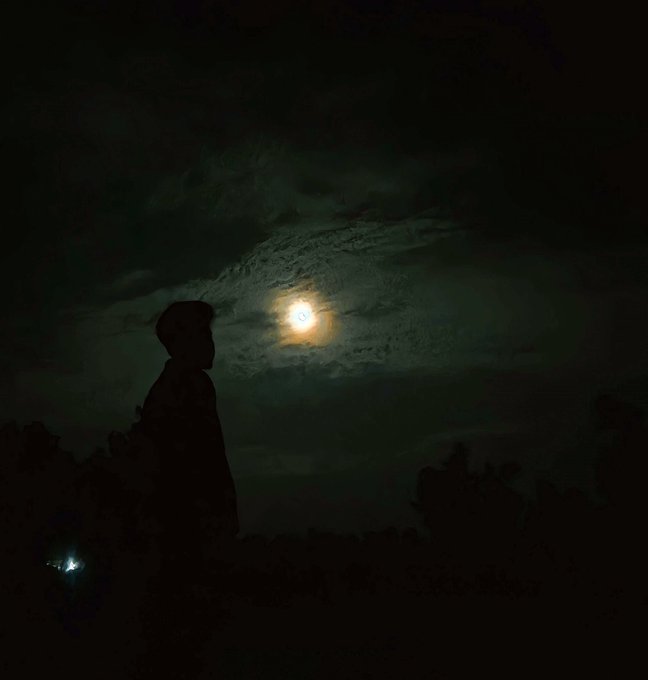 A dark nighttime photograph showing the silhouette of a person in profile against a cloudy sky illuminated by a bright full moon, with a small distant light visible in the lower left.