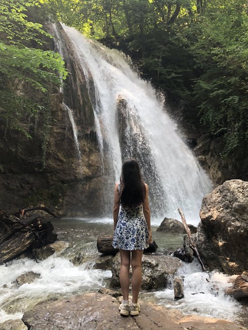 A woman with long dark hair in a blue floral dress stands on rocks at the base of a large waterfall cascading through a lush green forest.