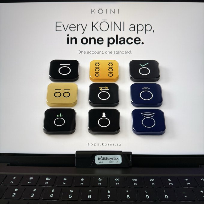 A display shows the KŌINI logo and 'Every KŌINI app, in one place. One account, one standard' above a 3x3 grid of physical button devices in black, yellow, and blue, with a KŌINIkeystick attached above a laptop keyboard.
