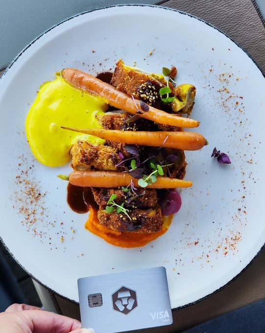 An overhead shot of a gourmet plated dish featuring roasted carrots, crusted protein pieces, yellow sauce, microgreens, and spice dusting on a white plate, with a Crypto.com Visa card held in the foreground.