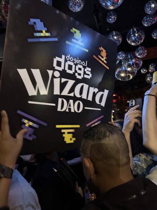 A nighttime concert/event photo showing a person holding up a sign that reads 'dosinal dogs Wizard DAO' decorated with Space Invaders-style pixel art, with disco balls visible in the background ceiling.