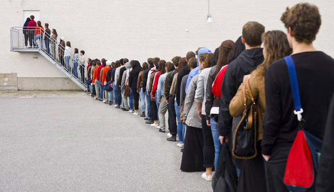 A long line of people waiting in a queue along a building with white brick walls.