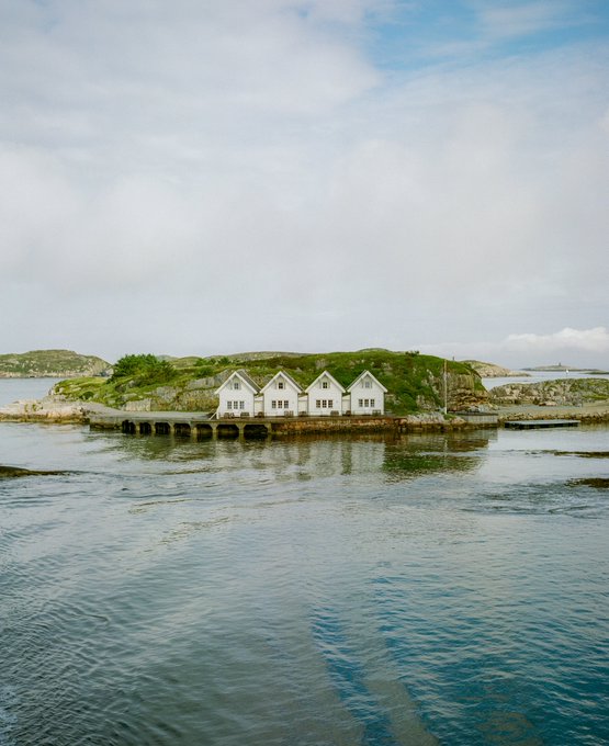 A small, grassy island in calm blue waters holds a row of identical white buildings with peaked roofs, sitting on a wooden pier structure with their reflections visible in the water below.