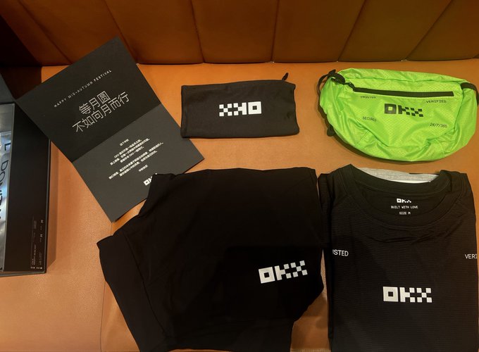 A collection of OKX branded merchandise, including black clothing and a green fanny pack, alongside a promotional booklet with Chinese text.