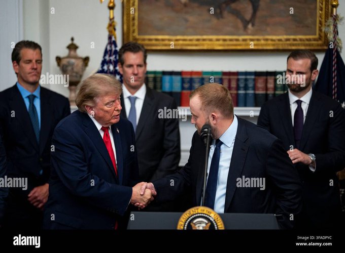 President Donald Trump shakes hands at a podium in the White House, as he stands beside another man in a dark suit, with three other men standing behind them, one of whom is Eric Trump. The image relates to the post's discussion of the Trump family's cryptocurrency dealings, specifically their receipt and transfer of LINK tokens to Coinbase.