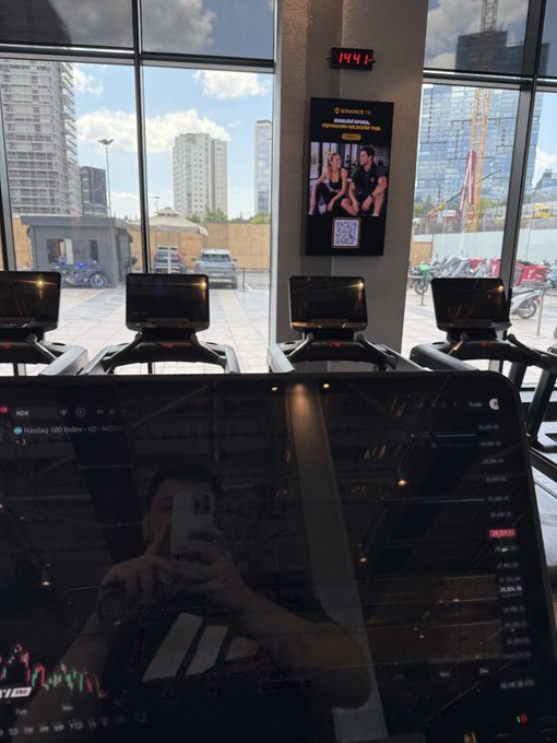 A person takes a mirror selfie on a treadmill while holding a phone, with a digital display showing stock/index charts (NDX, NDX1! 15m) reflecting back, and a Binance TR promotional screen on the wall announcing their partnership with MACTeam Runners. The image captures the post's theme of combining running with financial markets, now boosted by Binance TR's sponsorship of Turkey's largest running community of over 10,000 runners.