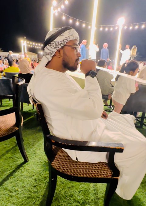 A man in traditional white Emirati attire with a keffiyeh and agal sits in a chair at an outdoor evening event, appearing to take a drink while watching the festivities. The string lights and gathering in the background suggest a social celebration, aligning with the post's theme of celebrating friends' successes.