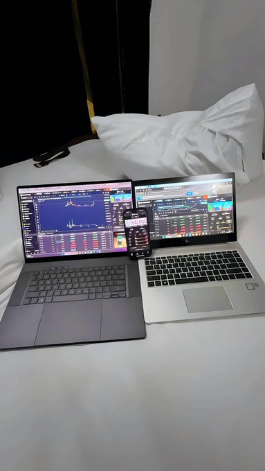 Two laptops and a smartphone displaying financial trading charts are set up on a white bed with pillows.