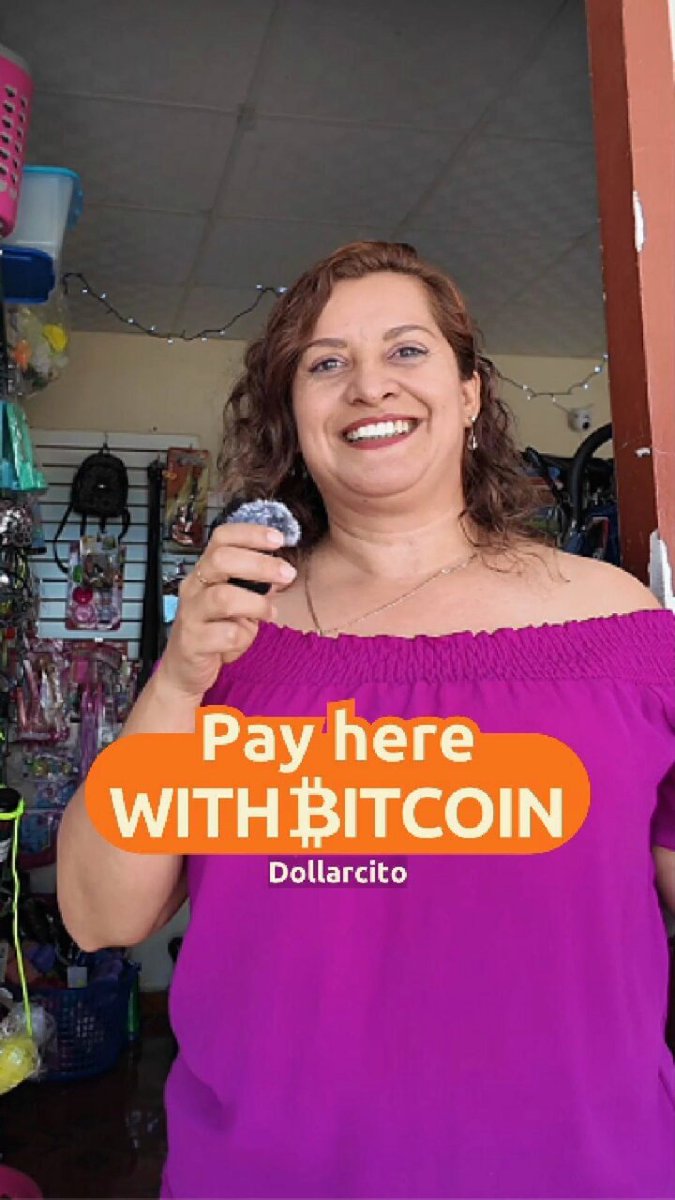 The image features a woman standing next to a door, holding a small object that appears to be a coin or token. She is wearing a purple top and smiling at the camera. The text "Pay Here with Bitcoin" is written above her head, indicating that she is accepting Bitcoin payments for goods and services in the area. There are also other people visible in the image, suggesting that this location might be a popular spot for locals or tourists to visit.
