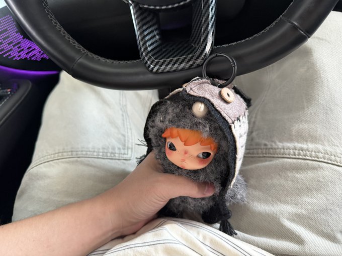 A hand holds a cute plush doll with orange hair and big eyes, wearing a fuzzy gray and pink hood with button details, hanging from a car steering wheel. The image relates to the post by showcasing the "毛茸茸的小野" (fuzzy little wild one) being affectionately presented, with purple ambient lighting visible in the car interior.