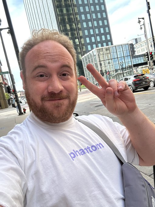 A man with a ginger beard takes a selfie outdoors on a city street, wearing a white 'phantom' t-shirt and flashing a peace sign.