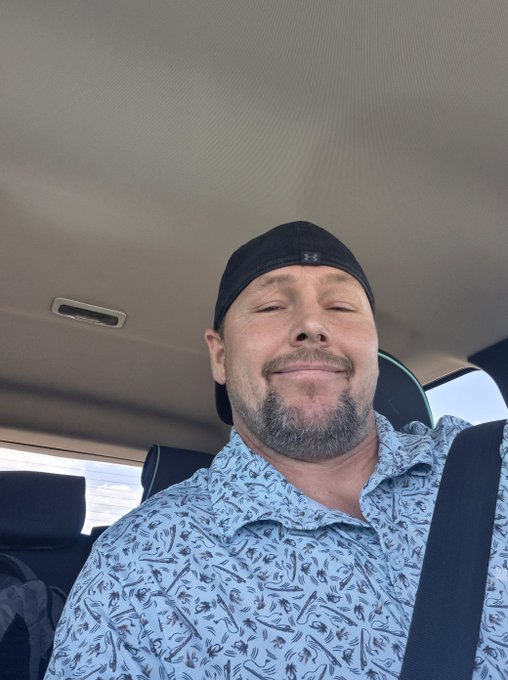 A bearded man wearing a black Under Armour cap and a light blue floral patterned shirt smiles for a selfie taken inside a vehicle.