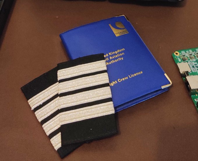 A pair of epaulettes with four gold stripes rests on top of a blue "United Kingdom Civil Aviation Authority Flight Crew Licence" booklet, displayed next to a circuit board on a brown surface.