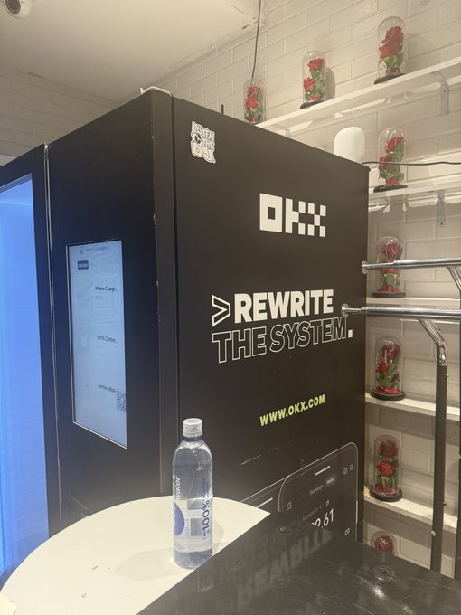 A black OKX cryptocurrency ATM/kiosk with the slogan 'REWRITE THE SYSTEM' and website 'www.okx.com' stands next to shelves displaying red roses under glass domes, with a water bottle on a round table in front.