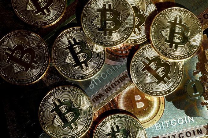 A close-up photograph of multiple gold-colored physical Bitcoin tokens stacked together, showing the Bitcoin 'B' symbol and the words 'BITCOIN' and '1 TROY OUNCE.'