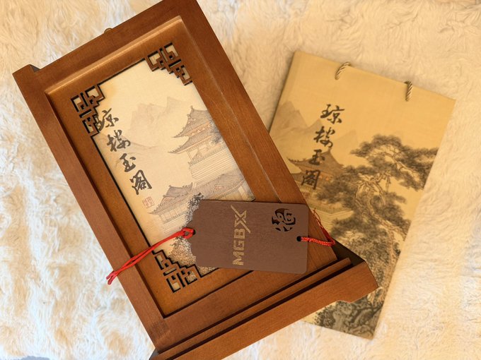 The image features a wooden box with a picture of a building, possibly a temple or pagoda, along with a tag that reads "MGBX" and "ZH". The box appears to be made out of wood and has a brown color. It is placed on top of a white surface, which contrasts nicely with the dark background. The image also includes text in Chinese characters, suggesting it might be related to Chinese culture or art.