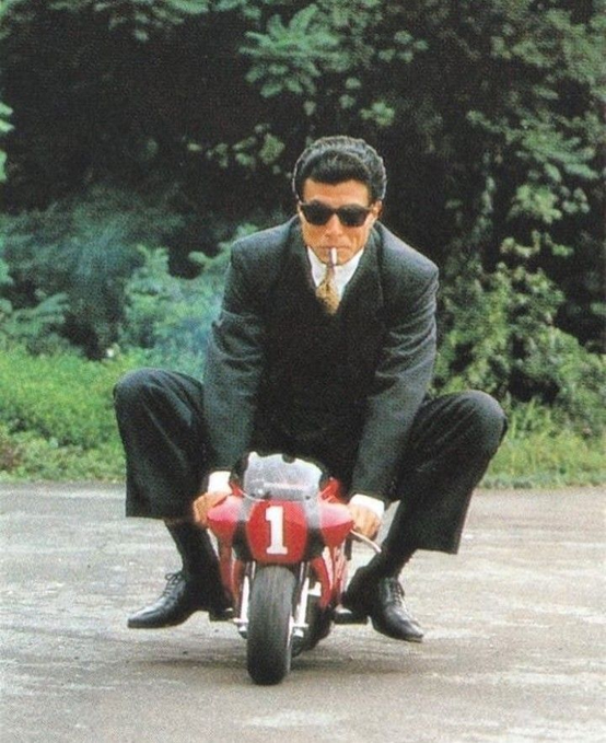 A man in a black suit, sunglasses, and tie sits crouched on a small red motorcycle marked with the number “1,” while holding a cigarette.