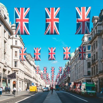 The image shows a London street scene, likely Regent Street, adorned with numerous Union Jack flags hanging above the road, symbolizing British identity and the Bank of England. This visual connects to the accompanying post about a Bank of England official commenting on stablecoins and dollar dominance, highlighting the tension between the U.S. dollar's 98% share of the stablecoin market and the UK perspective on this trend.