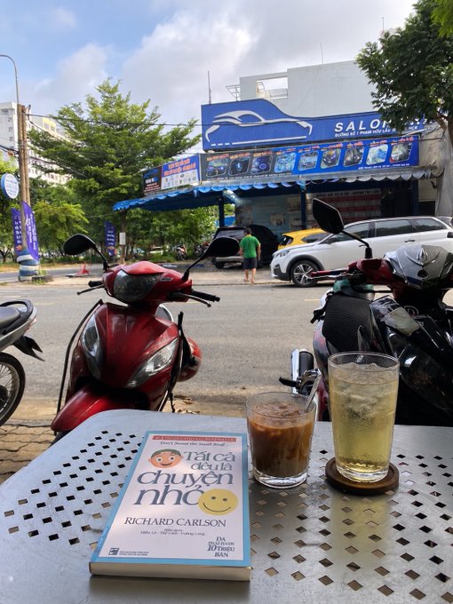 An outdoor café table at 'Salon' with two iced drinks and a Vietnamese book 'Tất cả đều là chuyện nhỏ' (Don't Sweat the Small Stuff) by Richard Carlson, with motorbikes parked on the street and a person walking in the background.