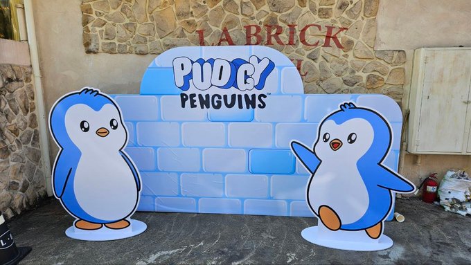 Two cardboard cutouts of blue and white cartoon penguins stand on either side of an inflatable light blue brick wall displaying the "PUDGY PENGUINS" logo, with "LA BRICK" visible on the wall behind.