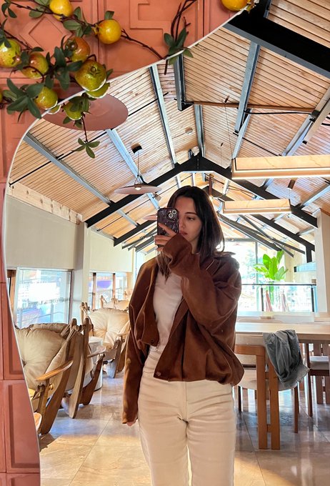 A woman takes a mirror selfie in a stylish café or restaurant, wearing a brown fleece jacket, white top, and white pants, with decorative oranges hanging above.