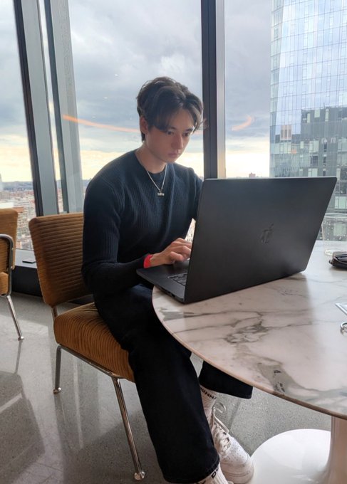 The image features a young man sitting at a table using his laptop computer. He appears to be focused on his work or possibly browsing the internet. The setting seems to be an office environment with chairs, a dining table, and a window providing natural light. There is also a cup placed near the edge of the table.
